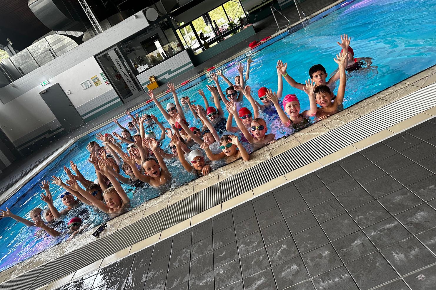 Mehrere Kinder mit Schwimmbrillen und Badekappen stehen im Wasser und heben die Hände in einem Schwimmbad.