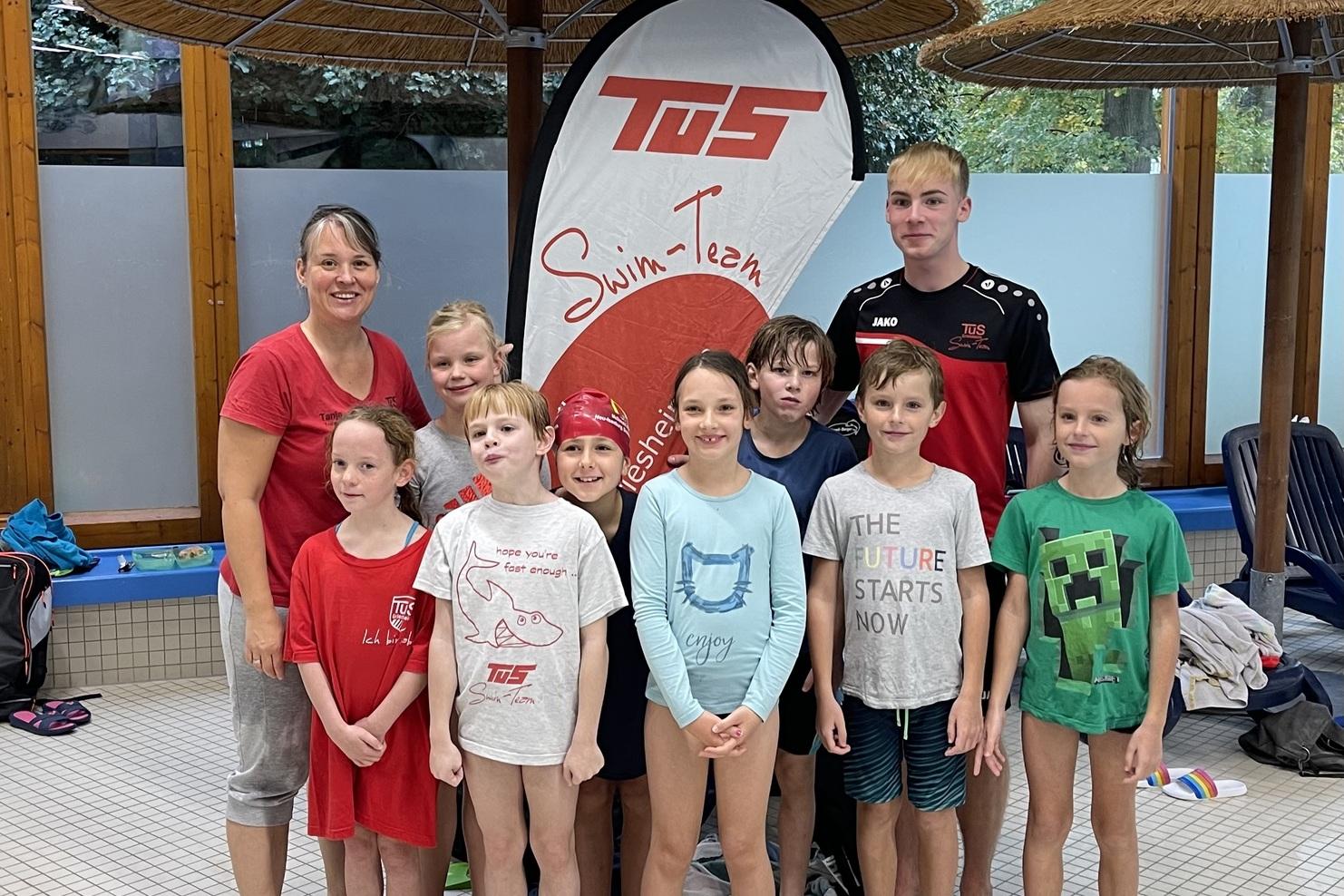 Gruppe von Kindern und Trainern vor einem Swim-Team-Schild in einem Hallenbad, umgeben von Wasser und Schwimmausrüstung.