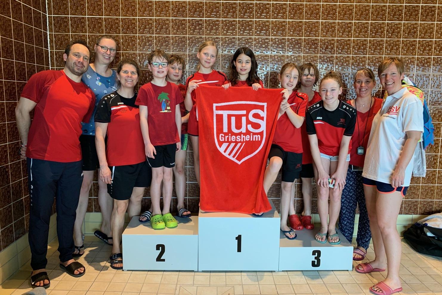 Gruppe von Schwimmern in roten Shirts posiert mit einer Medaille und einem Handtuch auf einem Siegerpodest.