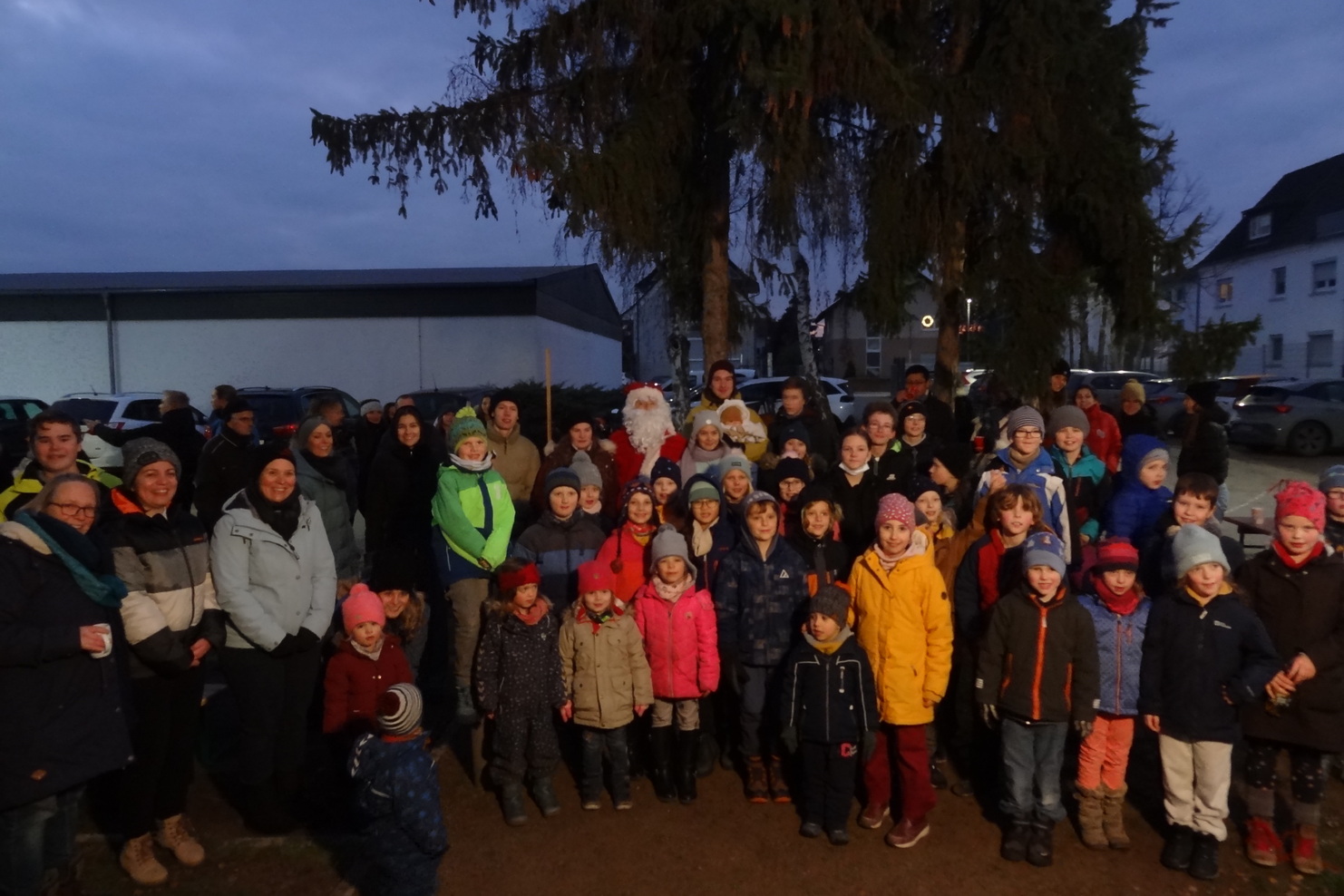 Gruppe von Kindern und Erwachsenen versammelt unter einem Baum bei Dämmerung, mit Weihnachtsmann in der Mitte.