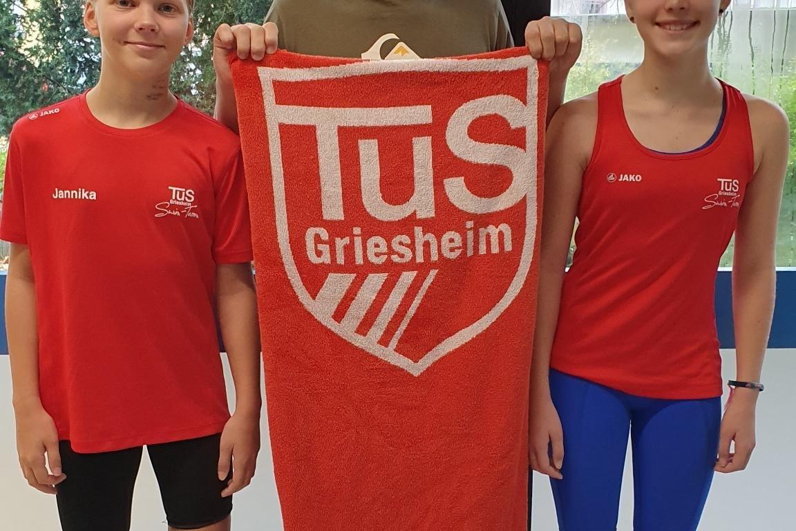 Drei Kinder in Sportkleidung stehen vor einem Fenster, einer hält ein Handtuch mit dem Logo von TuS Griesheim.