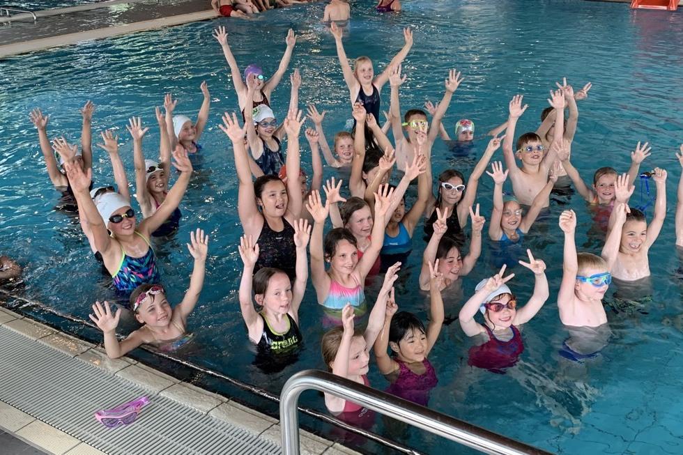 Gruppenaufnahme von Kindern im Schwimmbecken, die fröhlich die Arme heben und schwimmen.