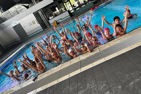 Mehrere Kinder mit Schwimmbrillen und Badekappen stehen im Wasser und heben die H&auml;nde in einem Schwimmbad.