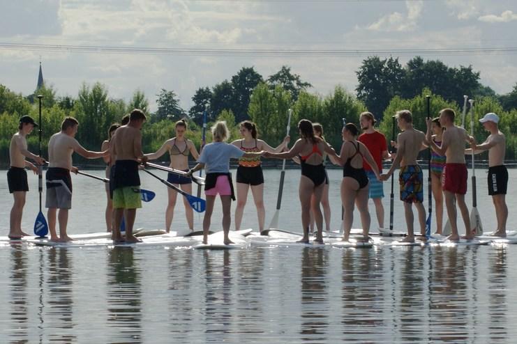 Gruppierung von jungen Erwachsenen, die auf Paddleboards stehen und sich gegenseitig halten, an einem ruhigen Gewässer.