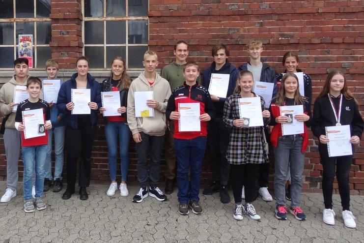 Gruppe von Jugendlichen posiert mit Urkunden und Medaillen vor einer Ziegelsteinmauer.