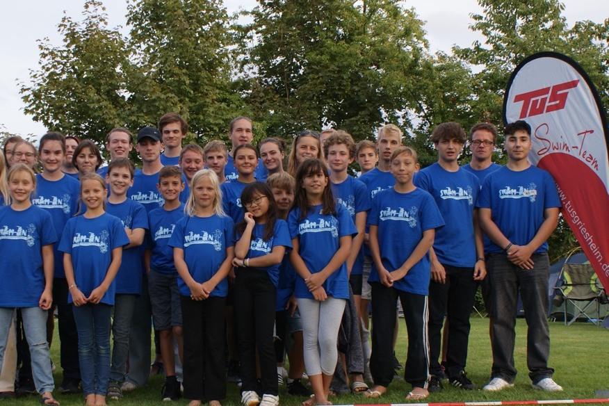 Gruppe von Kindern und Jugendlichen in blauen T-Shirts, versammelt auf einer Wiese vor B&auml;umen und einem Banner.