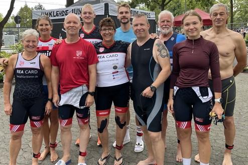 Gruppe von zehn Triathleten in Sportkleidung stehen auf einem Platz, lächelnd und bereit für ein Event.