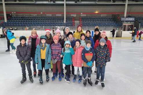 Gruppe von Kindern und Erwachsenen in Winterkleidung auf einer Eislauffläche, alle tragen Schlittschuhe.