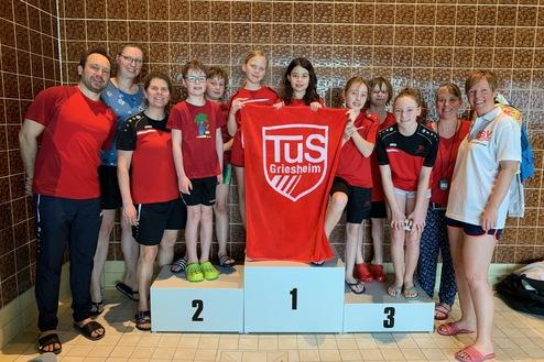 Gruppe von Schwimmern in roten Shirts posiert mit einer Medaille und einem Handtuch auf einem Siegerpodest.