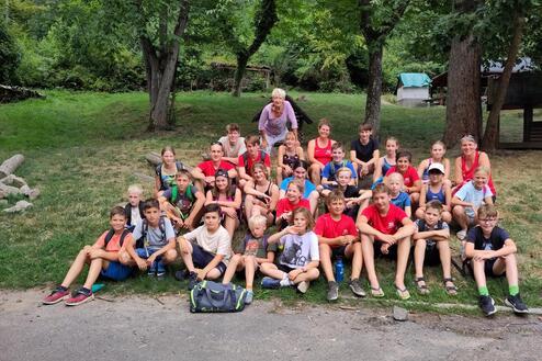 Gruppe von etwa 30 Kindern und Betreuern sitzend im Freien, umgeben von Bäumen und Wiese. Angenehme Sommersituation.