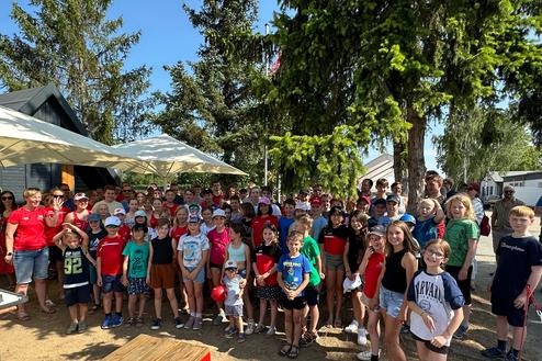 Gruppenszene mit Kindern und Jugendlichen im Freien, umgeben von Bäumen und Zelten, unter strahlend blauem Himmel.