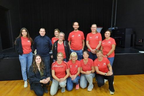 Gruppe von 12 Personen in roten T-Shirts, die lächeln und auf einer Bühne in einem hellen Raum stehen.