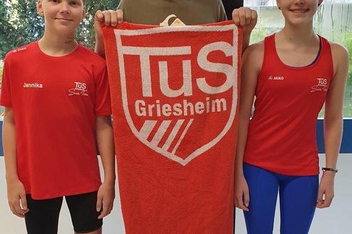 Drei Kinder in Sportkleidung stehen vor einem Fenster, einer hält ein Handtuch mit dem Logo von TuS Griesheim.