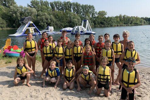 Gruppe von Kindern in Schwimmwesten am Strand, mit Wasserattraktionen im Hintergrund. Sonniger Tag am See.