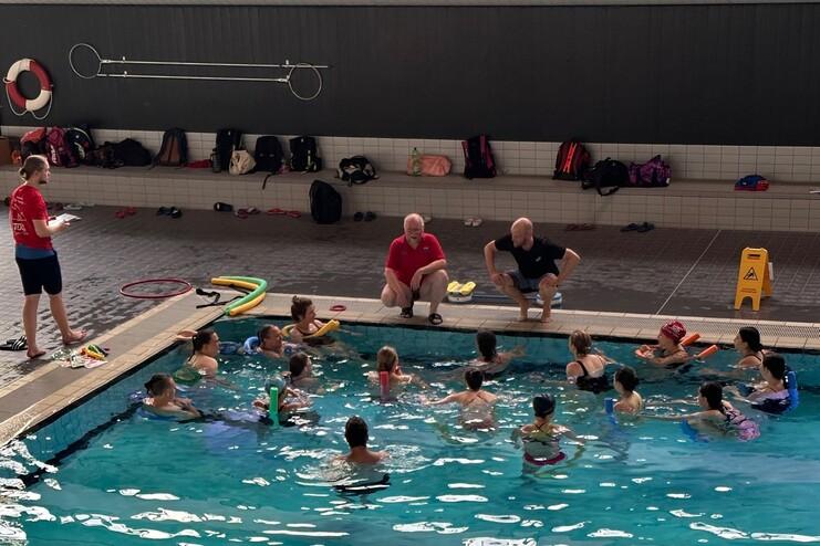 Gruppen von Kindern im Wasser mit Schwimmhilfen, während Trainer an Poolrand Anleitung geben. Schwimmutensilien auf dem Boden.