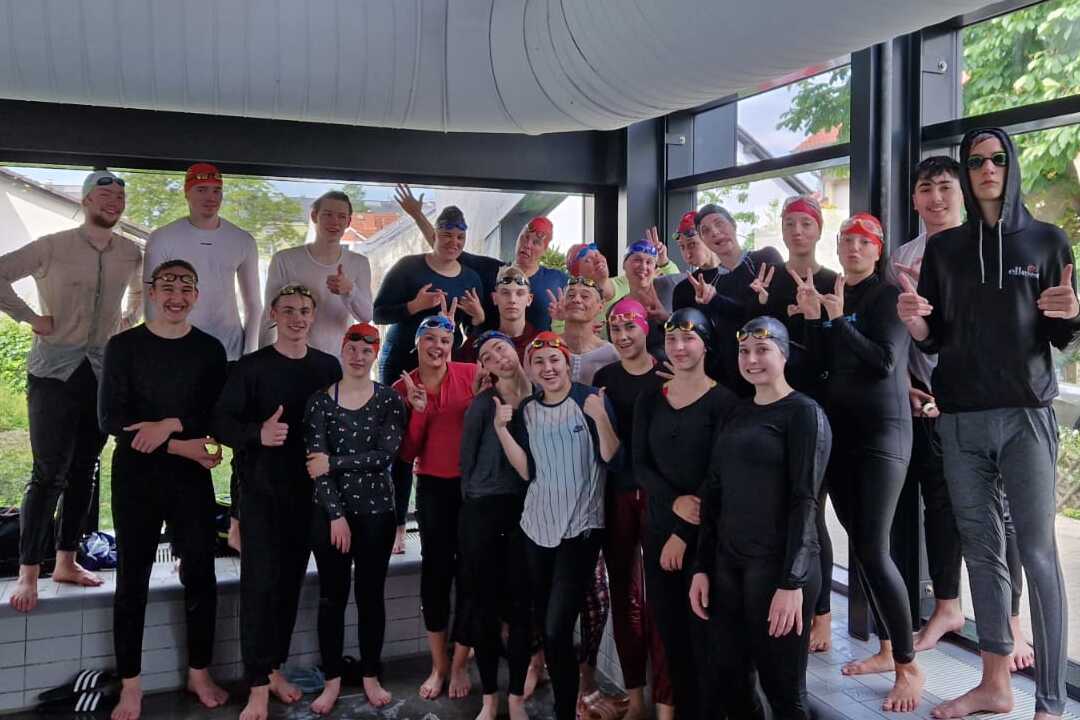 Gruppe von Schwimmern in Badebekleidung und Schwimmkappen posiert lächelnd vor großen Fenstern.