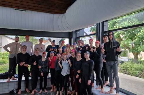 Gruppe von Schwimmern in Badebekleidung und Schwimmkappen posiert lächelnd vor großen Fenstern.
