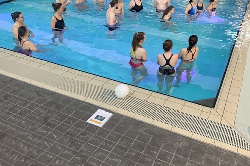 Gruppe von Personen steht im flachen Wasser eines Schwimmbeckens, umgeben von fliesenverkleideten Wänden.