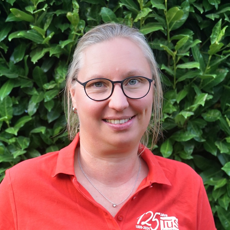 Frau mit Brille in rotem Polohemd steht vor einer grünen Hecke und lächelt freundlich in die Kamera.