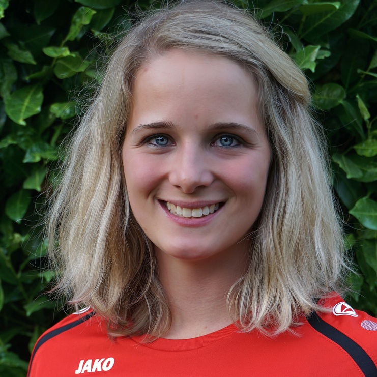 Porträt einer lächelnden Frau mit blonden Haaren in einem roten Sporttrikot vor einer grünen Pflanzenwand.