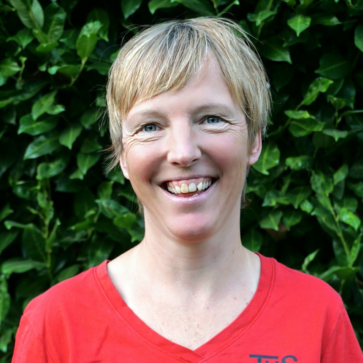 Porträt einer lachenden Frau mit kurzem Haar in rotem Shirt vor einer grünen Hecke.