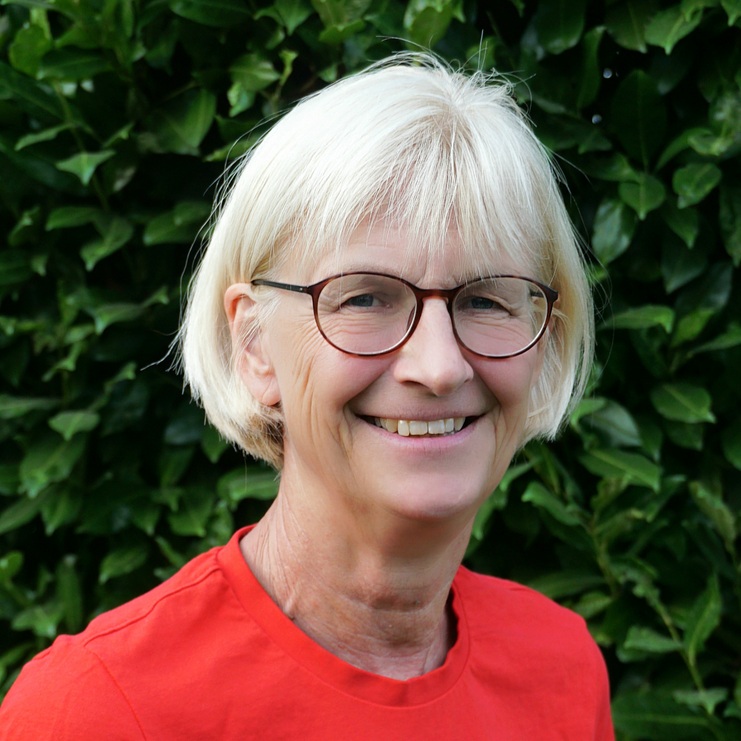 Lächelnde Frau mit blonden Haaren, trägt ein rotes T-Shirt mit Aufdruck, steht vor einer grünen Hecke.