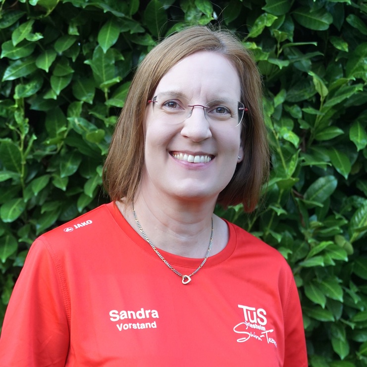 Porträt einer lächelnden Frau mit Brille, in rotem Sportshirt vor einer grünen Hecke.