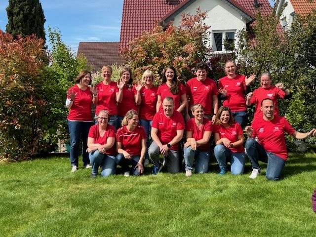 Gruppe von 14 Personen in roten T-Shirts, stehend und sitzend im Garten vor einem Haus, umgeben von grünen Pflanzen.