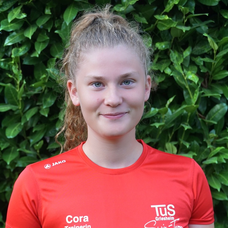 Porträt einer Frau in rotem Shirt mit Aufdruck, vor einer grünen Hecke stehend, lächelnd in die Kamera blickend.