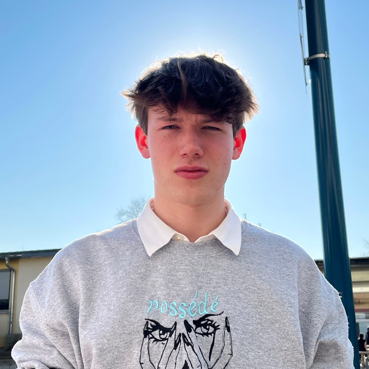Junger Mann mit braunen Haaren trägt ein graues Sweatshirt mit dem Schriftzug „possédé“ und einer grafischen Darstellung von Augen.