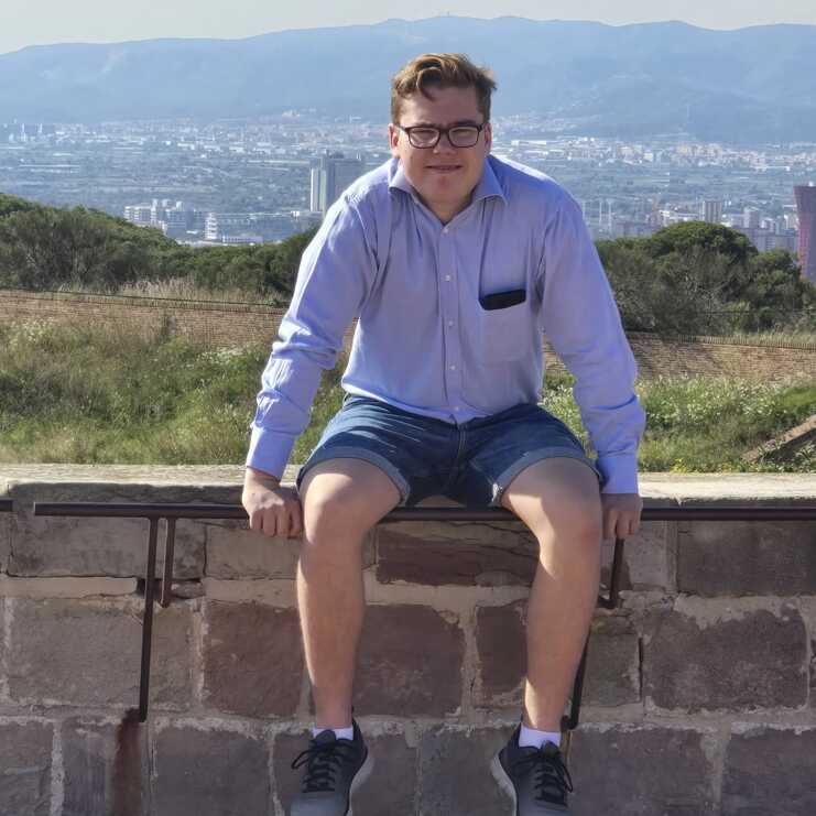 Junger Mann sitzt auf einer Steinmauer mit Blick auf eine Stadtlandschaft und Berge im Hintergrund.