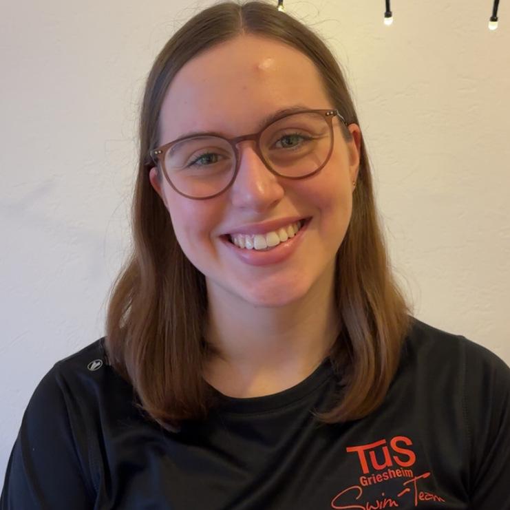 L&auml;chelnde Frau mit Brille tr&auml;gt ein schwarzes Shirt mit dem Logo des TuS Griesheim Swim Teams. Hintergrund mit Lichterkette.