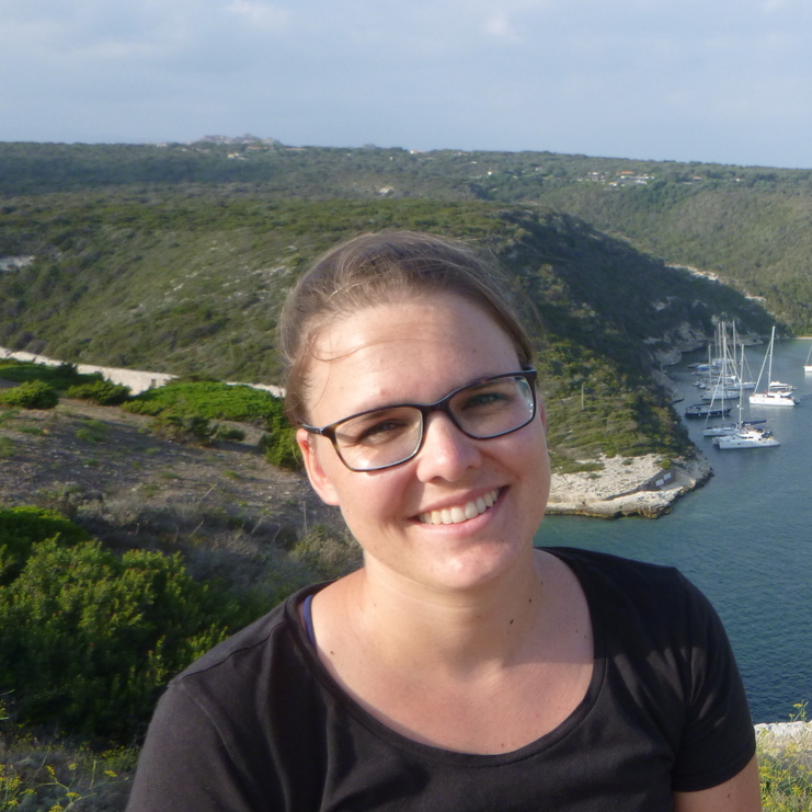 Frau mit Brille lächelt vor einer Küstenlandschaft mit Yachten im Wasser und bewachsenen Hügeln.