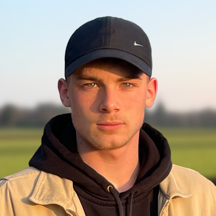 Junger Mann in einem beigen Jackett und schwarzem Hoodie steht vor einer grünen Wiese bei Sonnenuntergang.