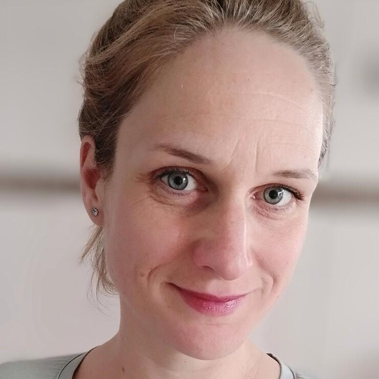 Porträt einer lächelnden Frau mit blauen Augen, die ein hellgrünes Sweatshirt trägt. Hintergrund unscharf.