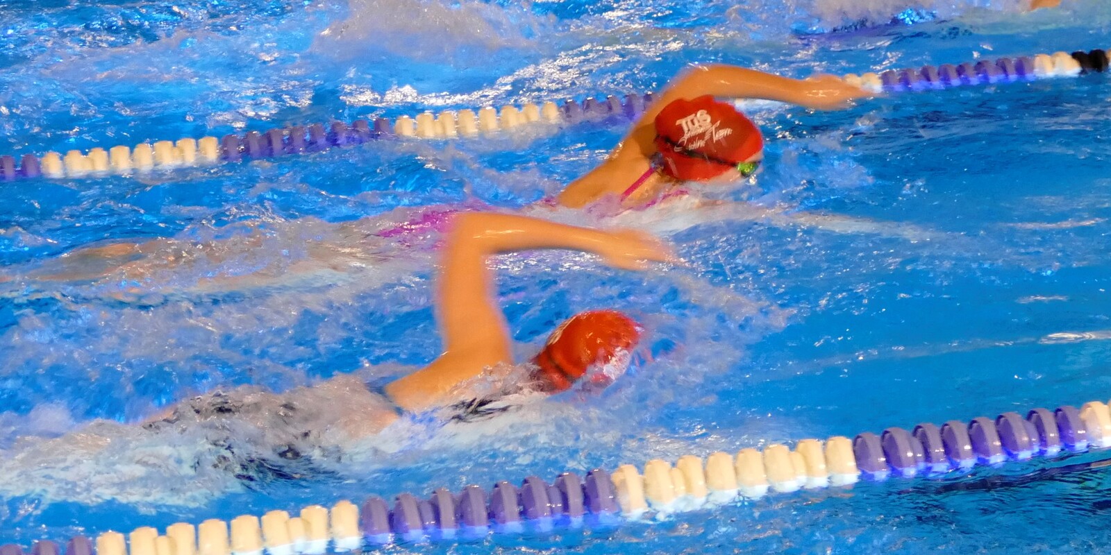 Zwei Schwimmerinnen in roten Badekappen schwimmen im Wettkampf in einem blauen Schwimmbecken zwischen Bahnmarkierungen.