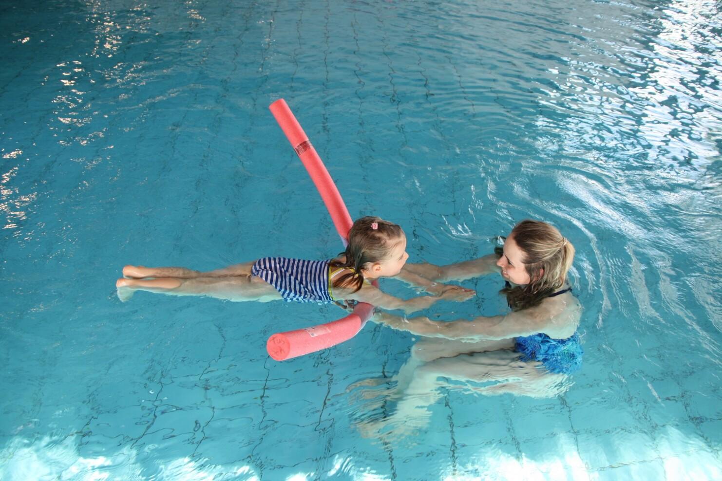 Zwei M&auml;dchen im Wasser, das kleinere nutzt einen Schwimmnudel, w&auml;hrend das andere sie h&auml;lt und anl&auml;chelt.