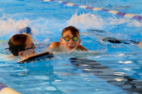 Zwei Kinder mit Schwimmbrillen spielen im Wasser eines Schwimmbeckens, einer hält ein Schwimmbrett.