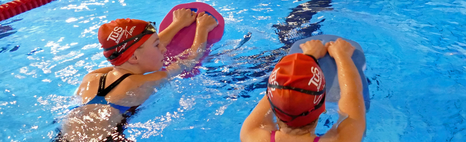 Zwei Schwimmerinnen mit Badekappen &uuml;ben im Wasser mit Schwimmbrettern in einem Hallenbad.