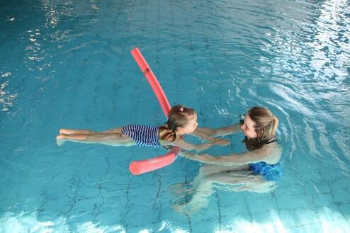 Zwei M&auml;dchen im Wasser, das kleinere nutzt einen Schwimmnudel, w&auml;hrend das andere sie h&auml;lt und anl&auml;chelt.
