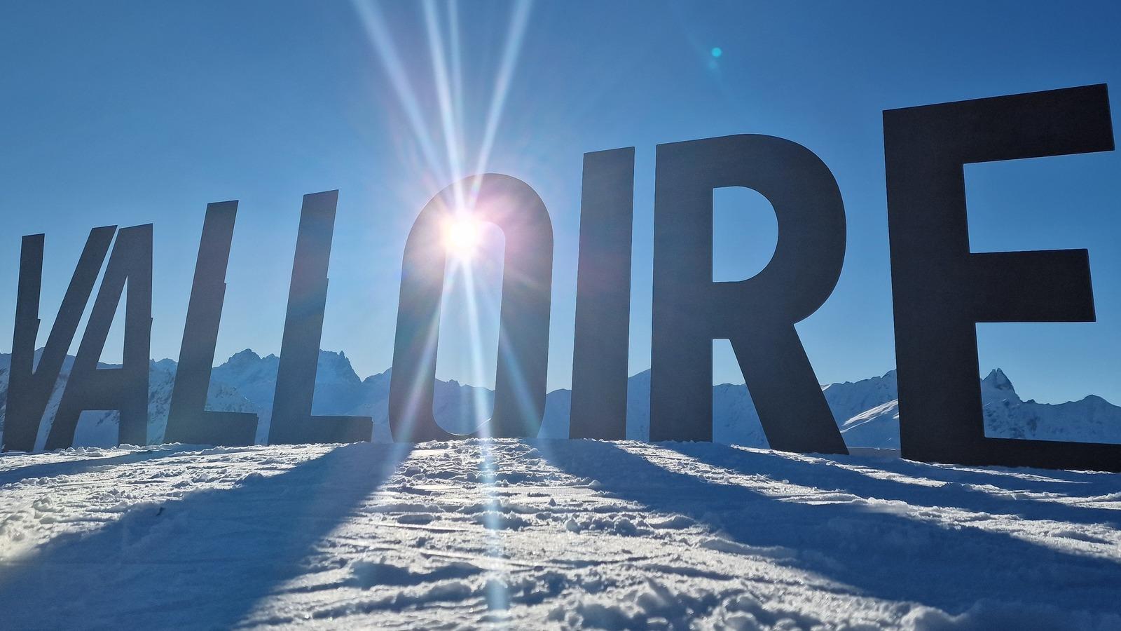 Sonnenstrahlen hinter dem Schriftzug "VALLOIRE" auf schneebedecktem Boden vor Bergpanorama.