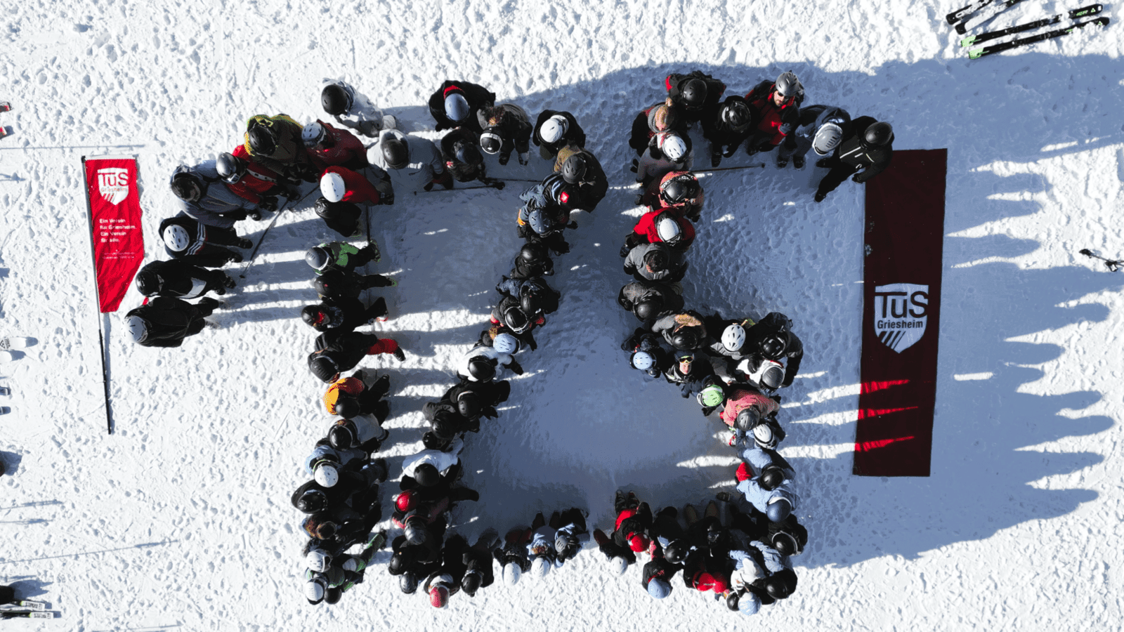 Gruppierung von Personen auf einem verschneiten Hang, die gemeinsam ein gro&szlig;es "T" formen, mit einem roten Banner im Hintergrund.