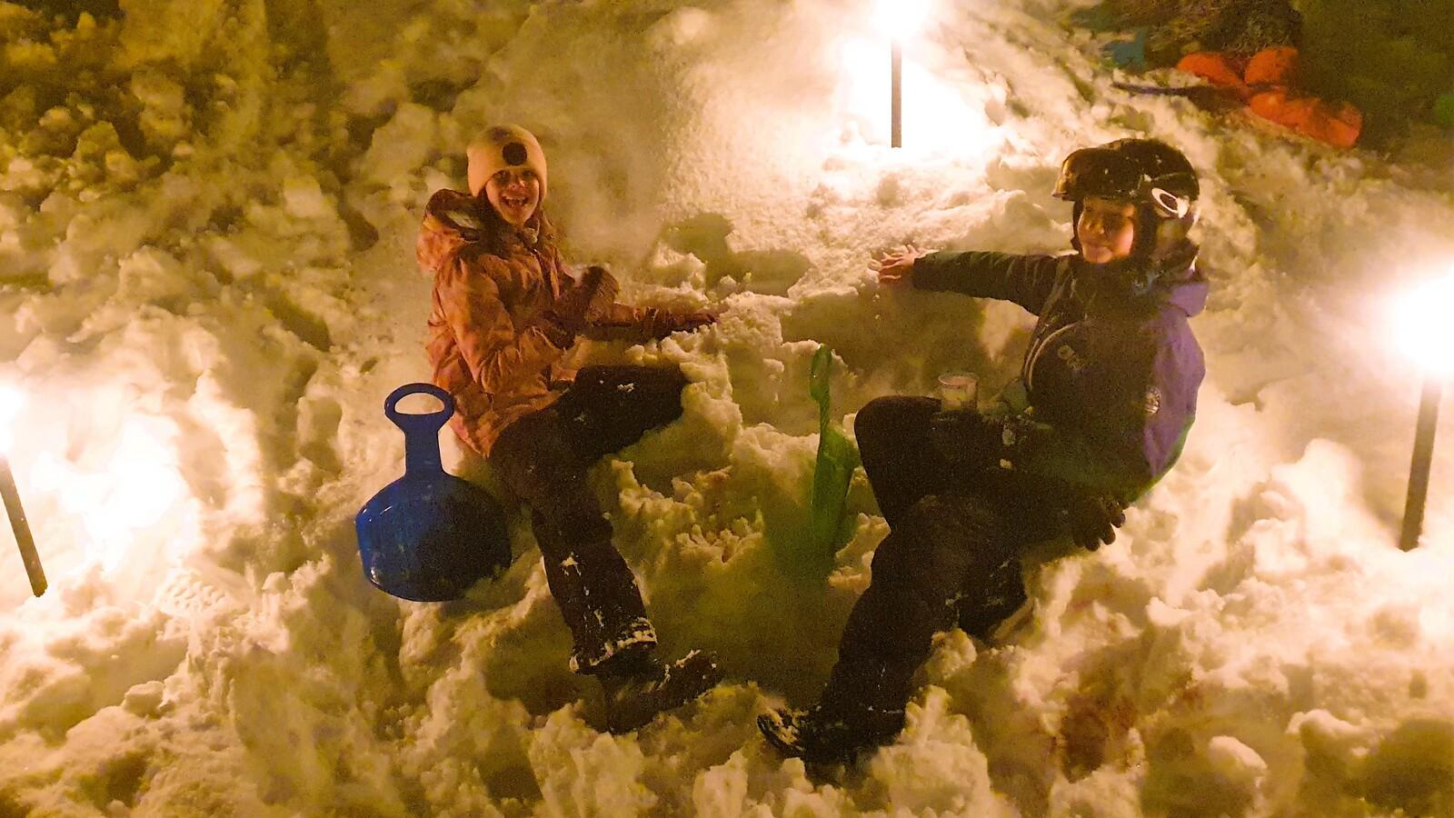 Zwei Kinder sitzen in einem Schneehaufen, umgeben von Fackeln, mit einem blauen Schlitten und einem gr&uuml;nen Spielzeug.