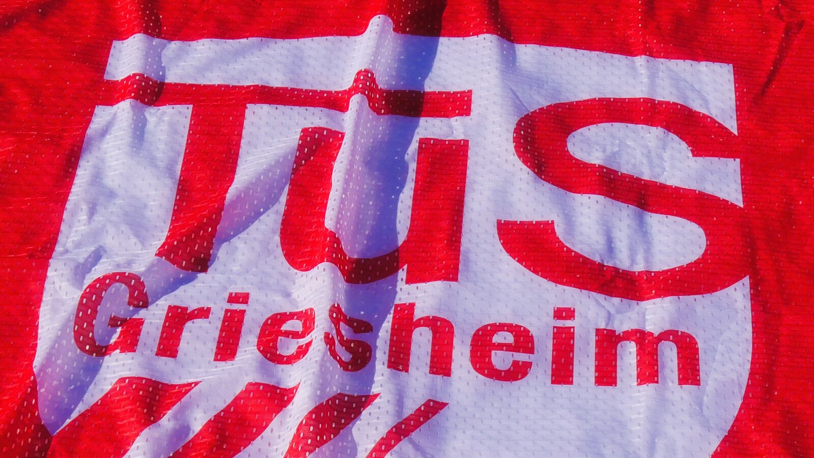 Logo des TuS Griesheim auf rotem Hintergrund, mit klarer, wei&szlig;er Schrift und Wellenmuster.