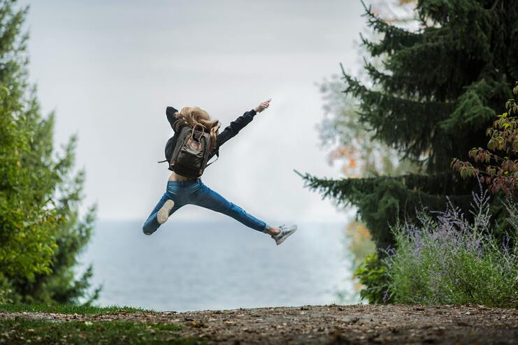 Junge Person springt fr&ouml;hlich mit Rucksack auf einem Weg mit Blick auf einen See und umliegende B&auml;ume.