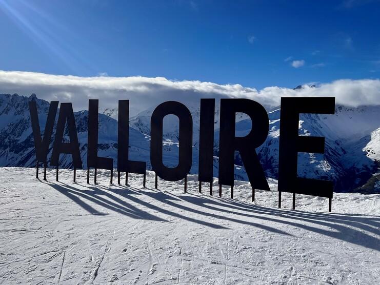 Schild mit der Aufschrift „VALLOIRE“ steht auf schneebedecktem Boden, umgeben von Berglandschaft und blauem Himmel.