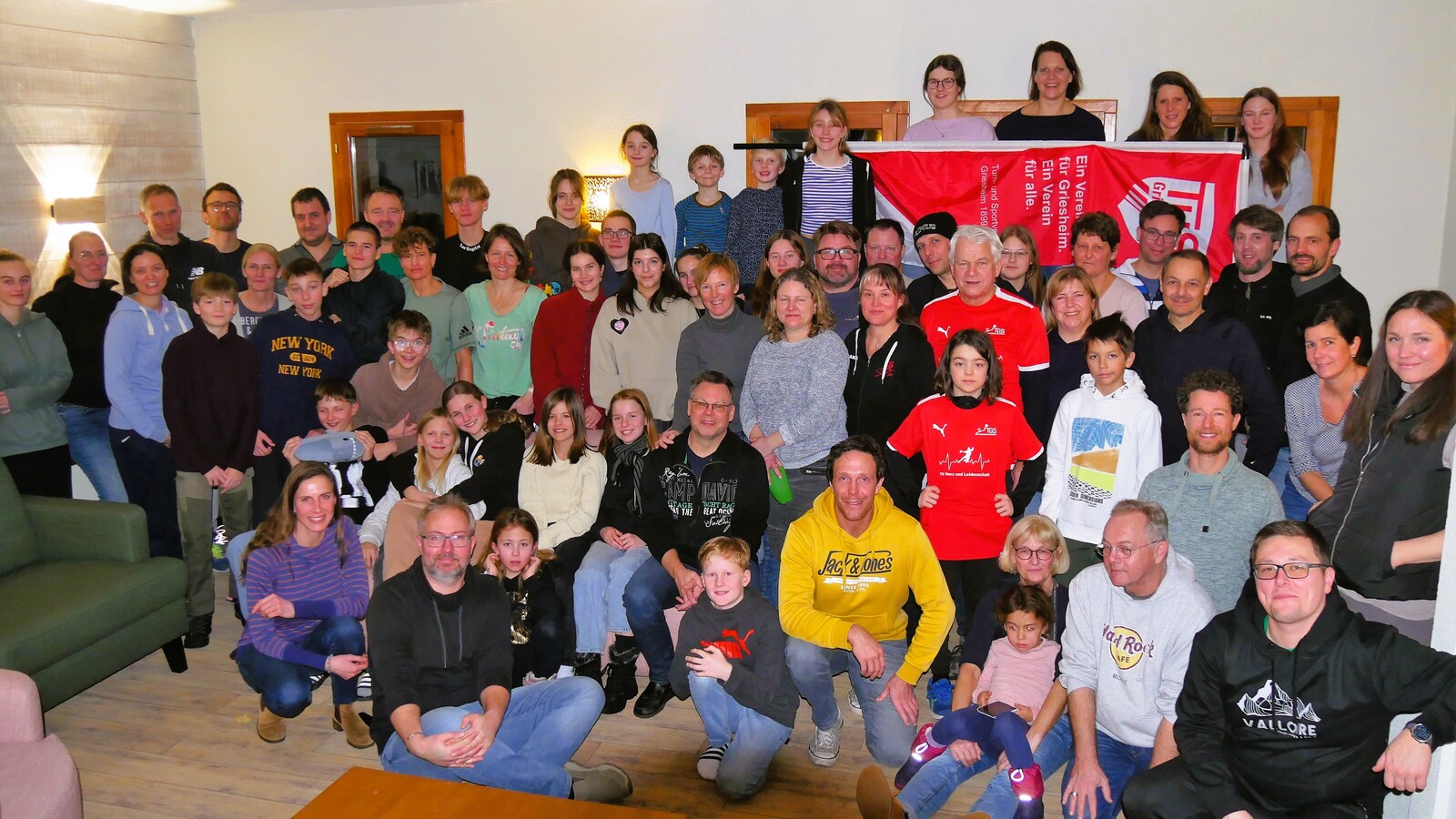 Gruppenfoto mit etwa 60 Personen in einem Raum, einige sitzen, andere stehen vor einer roten Vereinsflagge.