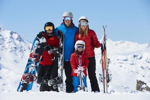 Familie posiert in Skioutfit im Schnee, zwei Kinder halten Snowboards, Erwachsene halten Skier, schneebedeckte Berge im Hintergrund.