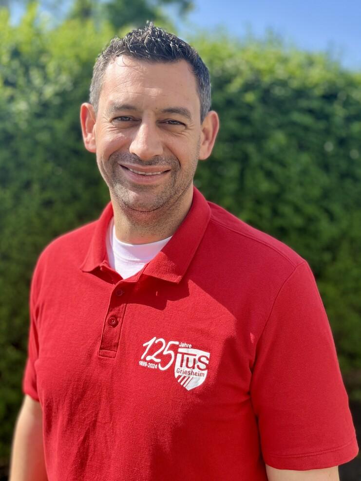Lächelnder Mann im roten Poloshirt mit Vereinslogo vor einer grünen Hecke und blauem Himmel.