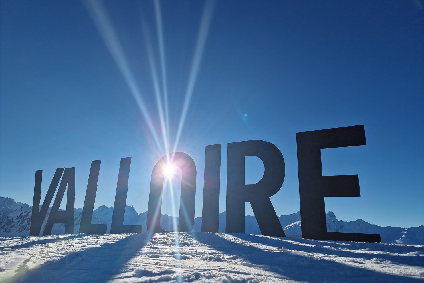Schneebedeckter Boden mit dem Schriftzug "VALLOIRE" vor einem klaren blauen Himmel und schneebedeckten Bergen im Hintergrund.
